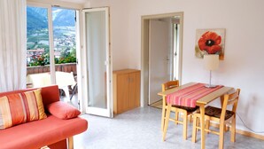 A room with a red sofa, a wooden dining table with four chairs, a cabinet, and a large window offering a view of a town and mountains. - Apartment Weitblick - Alpstay (Naturno)