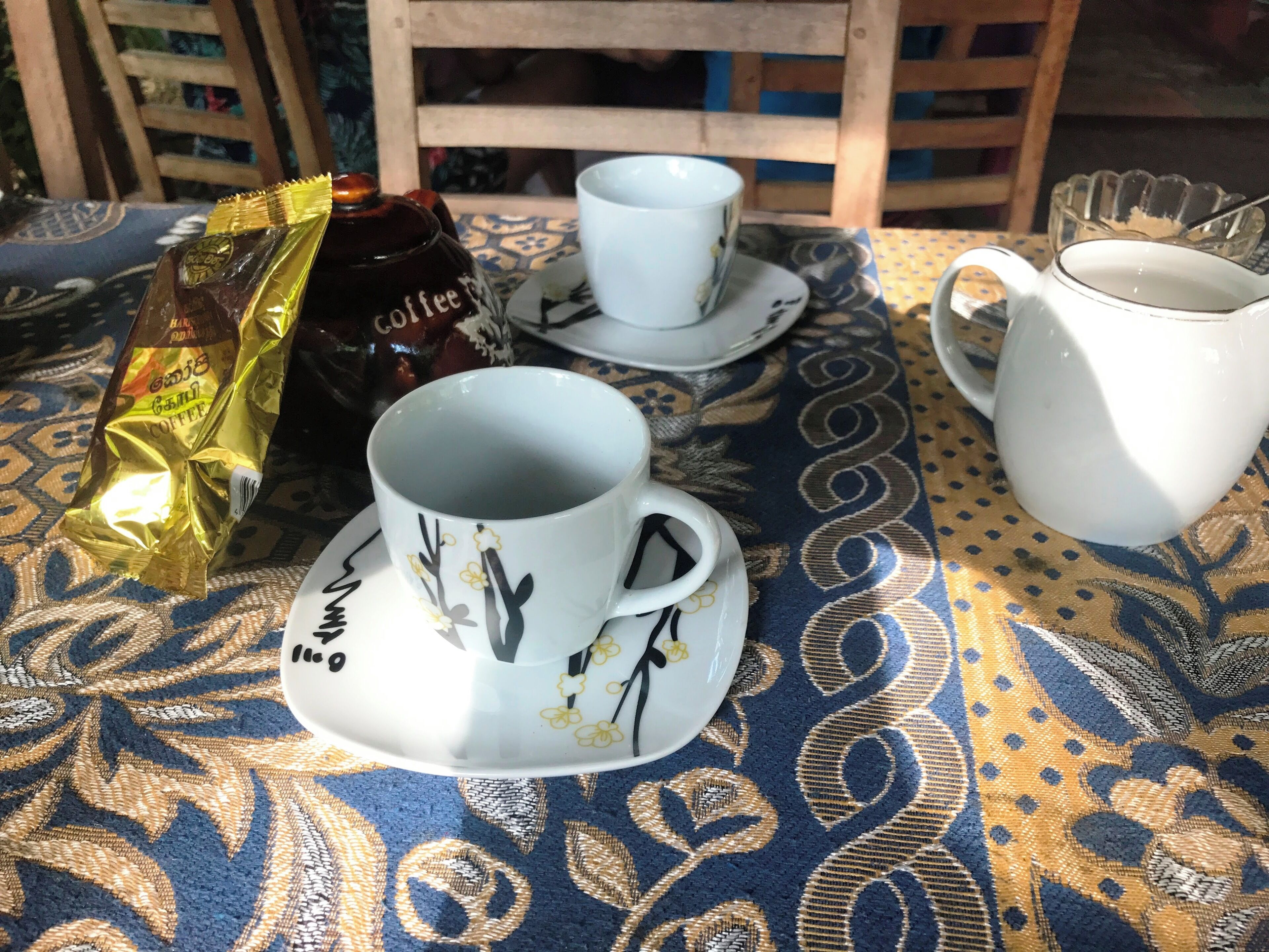 A table with a patterned tablecloth, a coffee bag, two cups, and a pitcher.