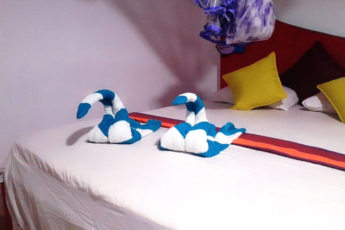 A bed with a white bedspread and two decorative swan figures made of fabric. There are pillows in yellow and red colors, and a purple floral item hanging on the wall.