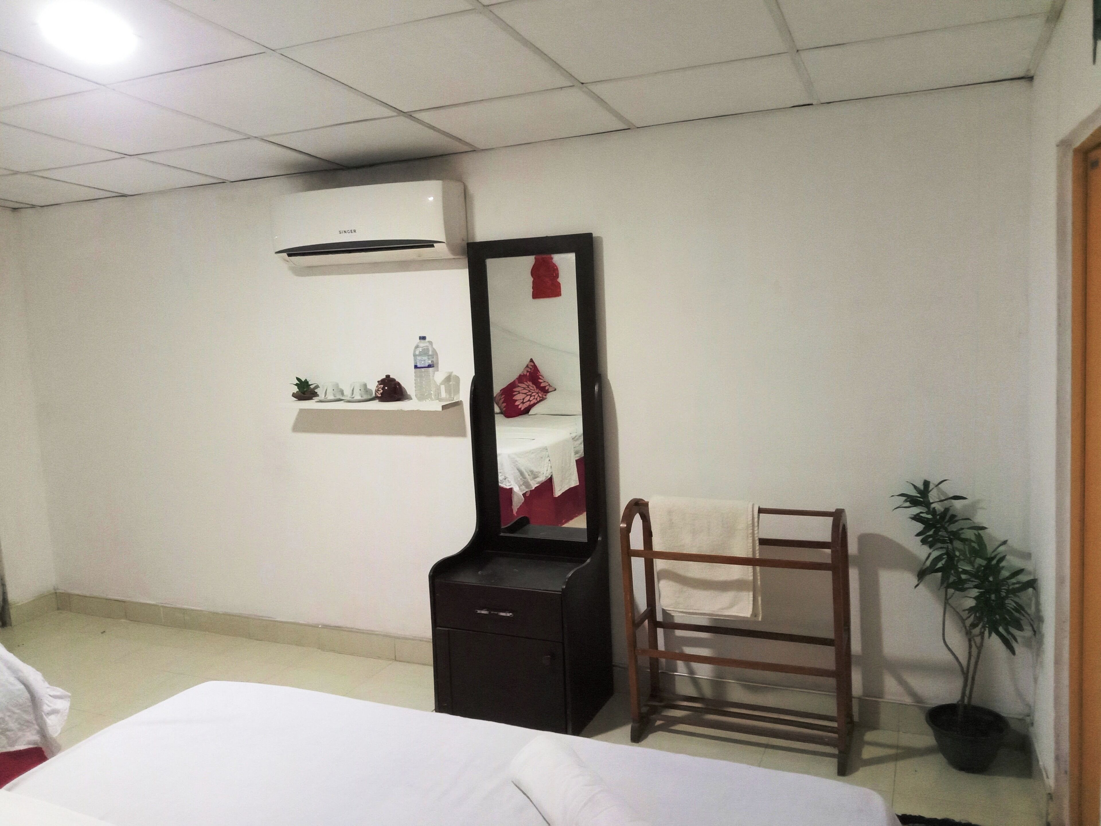 A hotel room with a bed, a mirror, a shelf with toiletries, a towel rack, and a potted plant.
