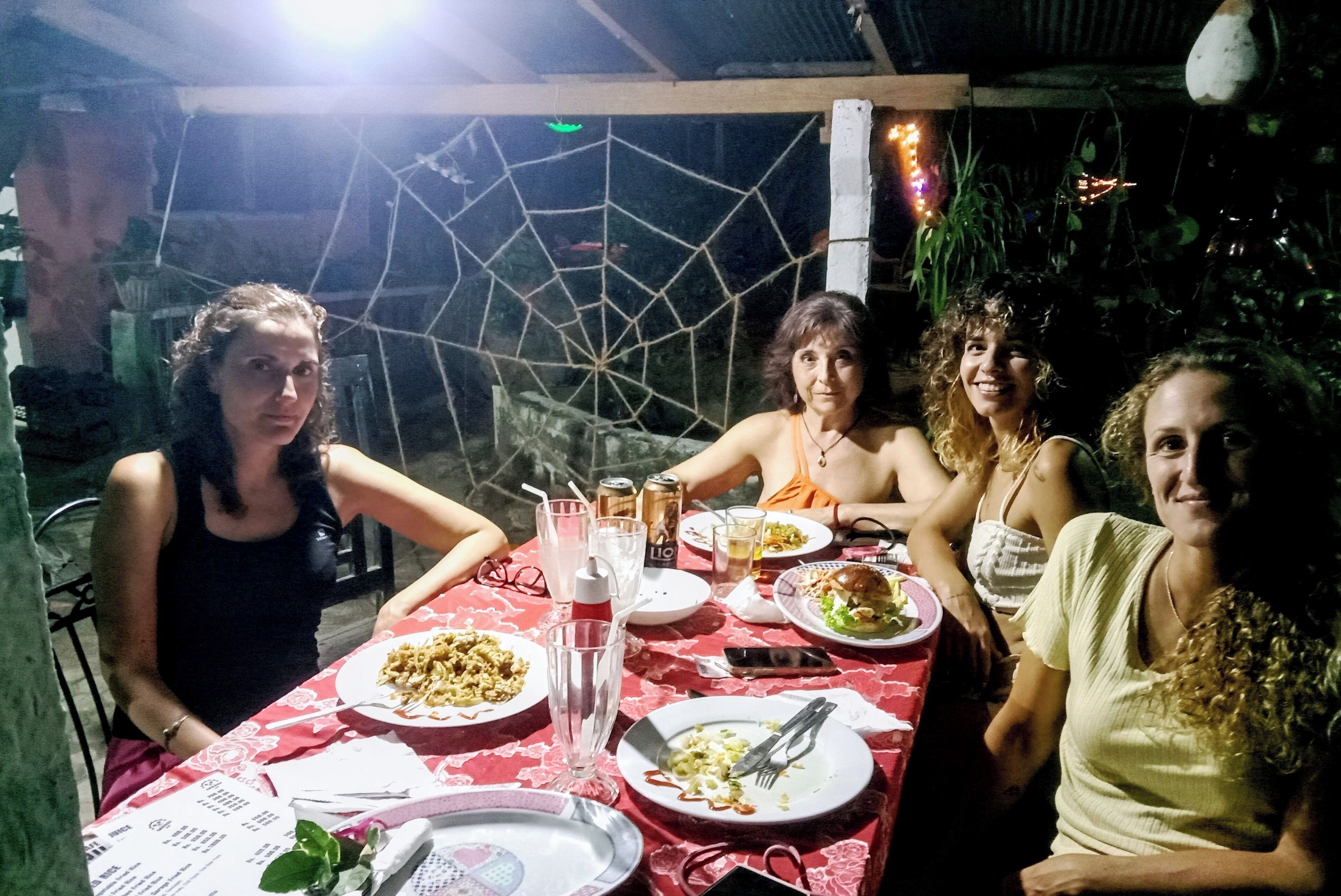A group of people dining at an outdoor table with a red tablecloth, surrounded by plants and a decorative spider web.