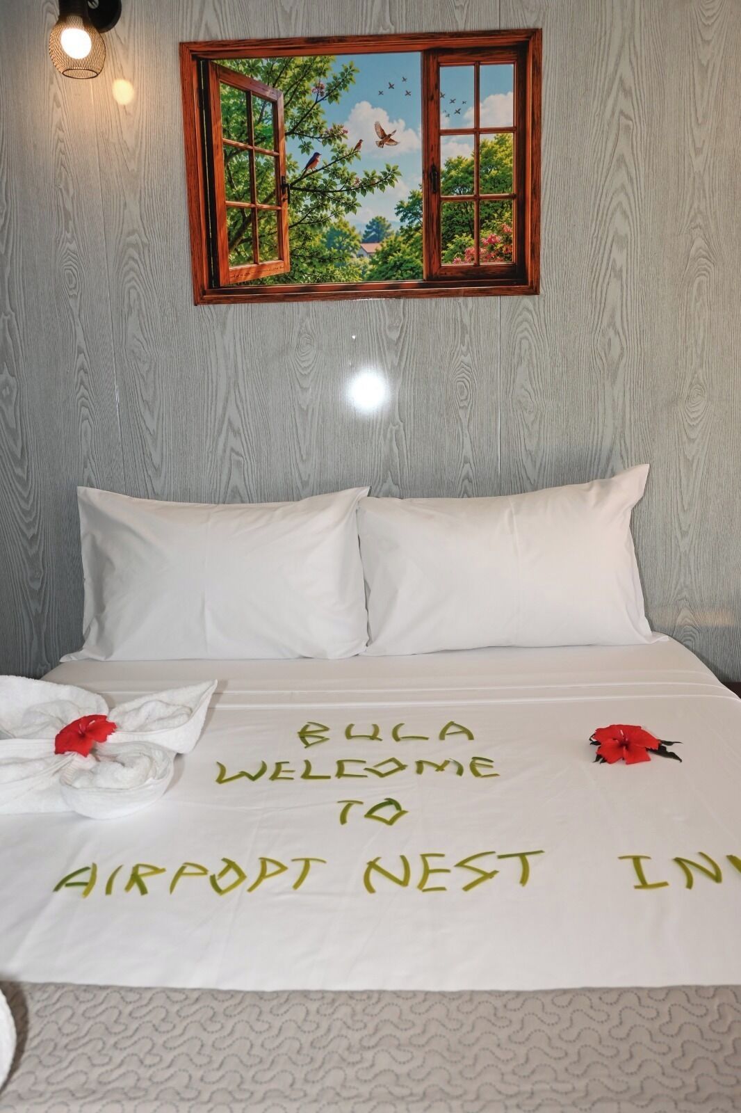 A neatly made bed with a welcome message, a window with a view of trees and birds, and a wall-mounted light fixture.