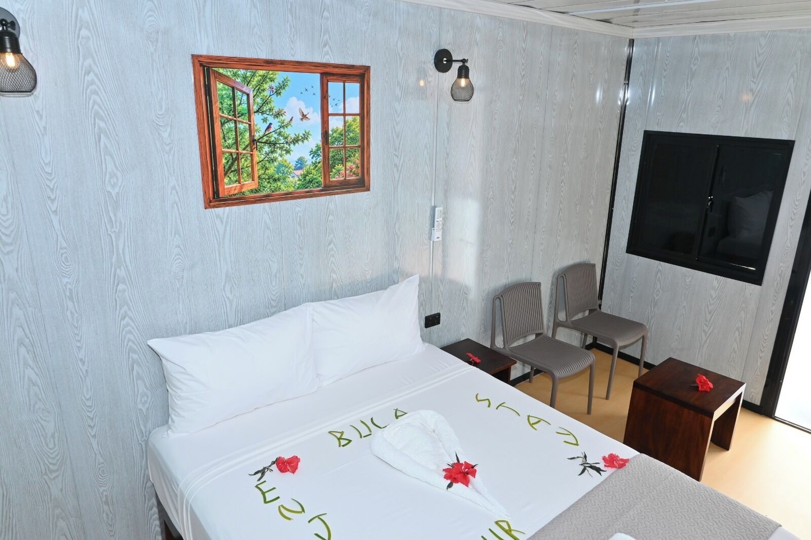 A hotel room with a bed, a desk, two chairs, a TV, and a wall-mounted picture of a window with trees and birds.