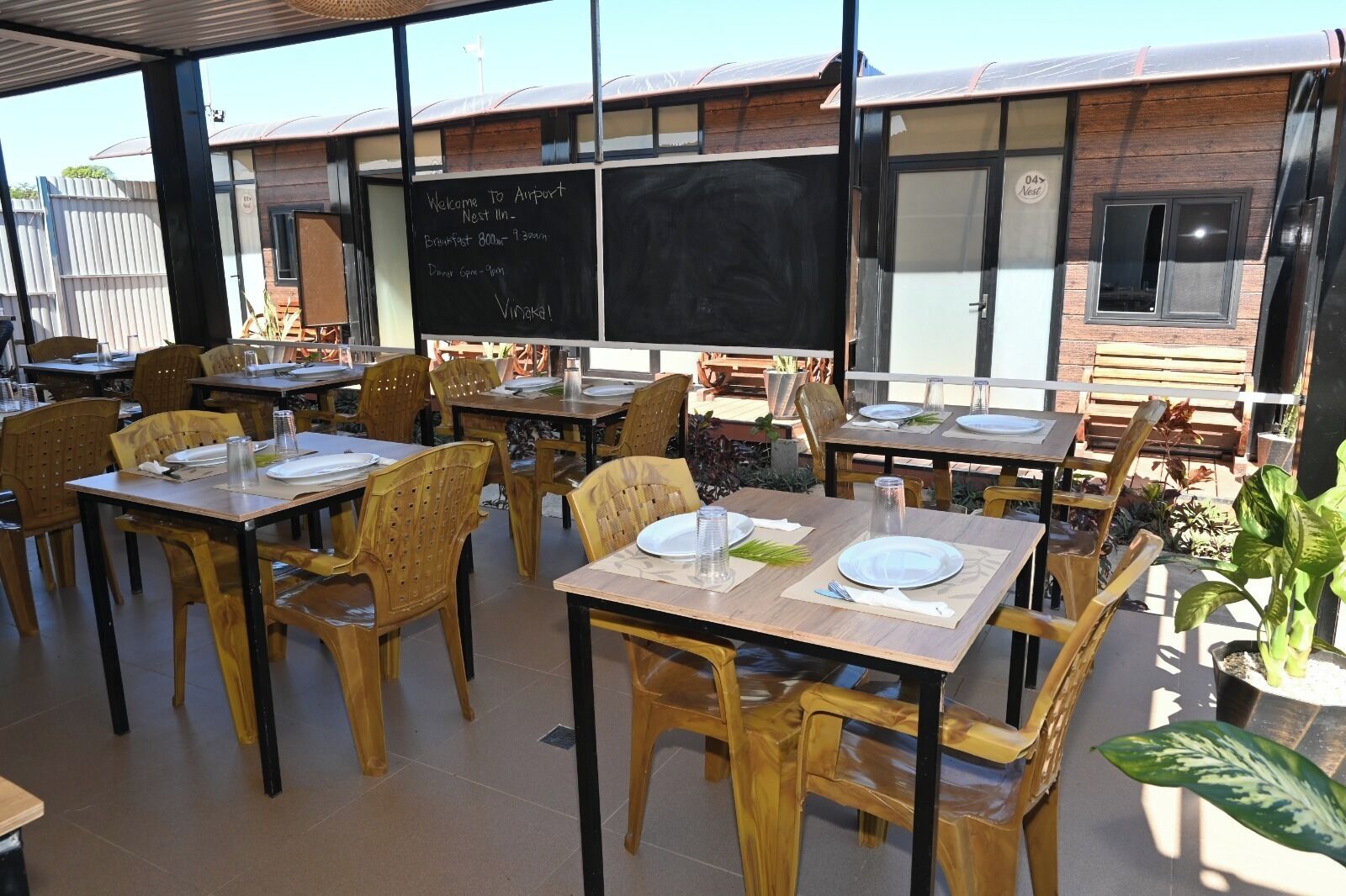 An outdoor dining area with tables and chairs, a blackboard with information, and a building with a wooden facade in the background.