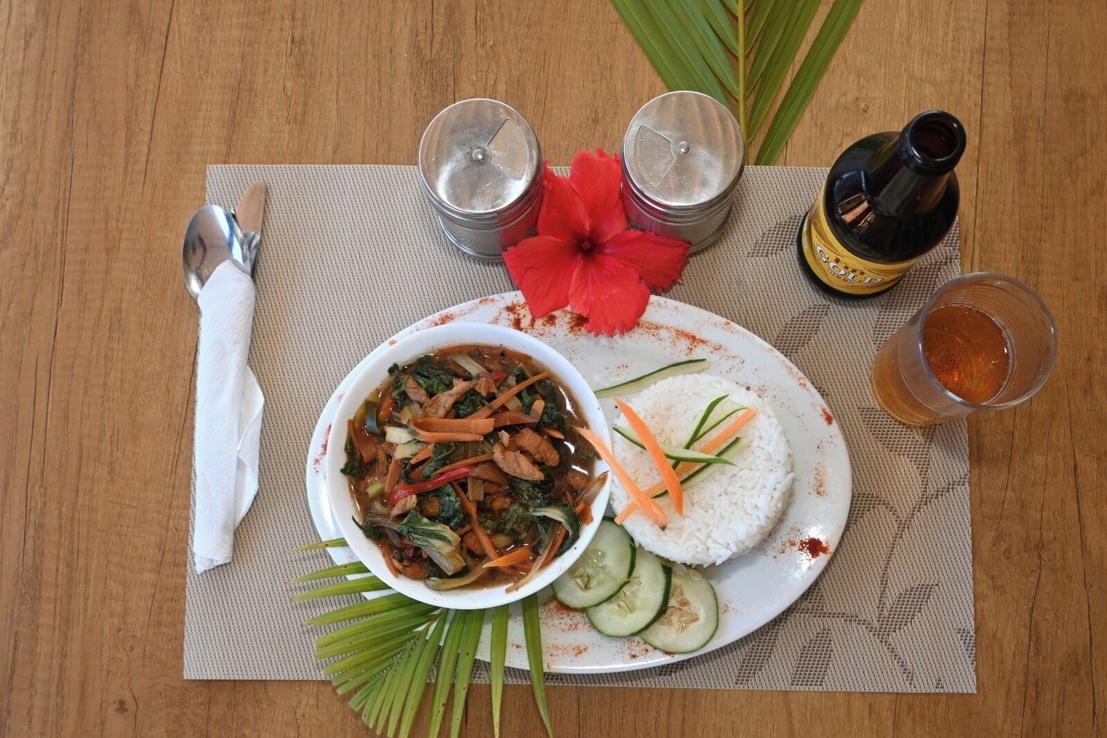 Comida con arroz, verduras y carne, una botella de salsa, un vaso de bebida y una flor roja sobre una mesa.