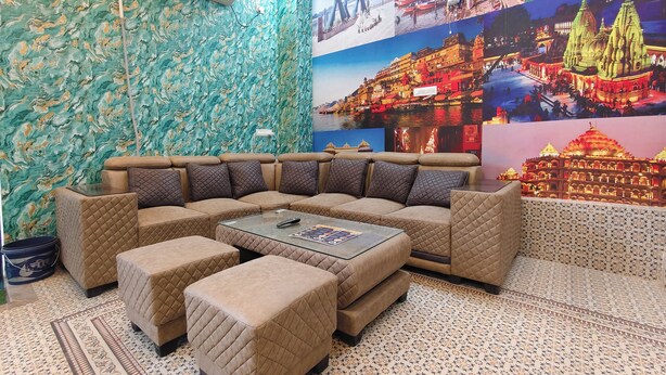 A living room with a sectional sofa, ottomans, a glass coffee table, and a wall decorated with travel posters. - Hotel Shiv Sagar  (Varanasi)