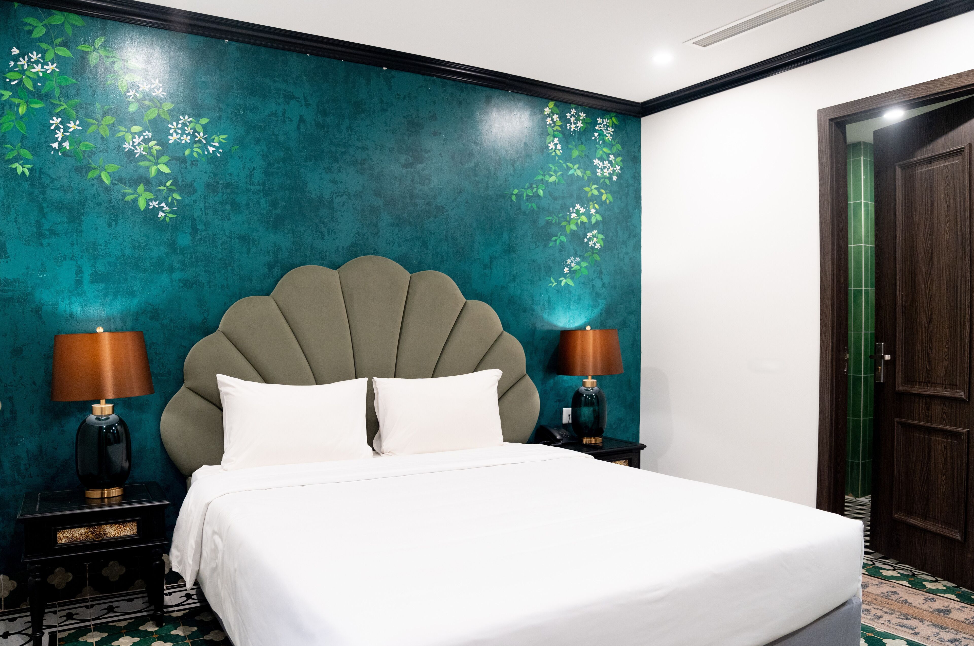 A neatly made bed with white linens, two bedside lamps, and a teal textured wall with floral patterns.