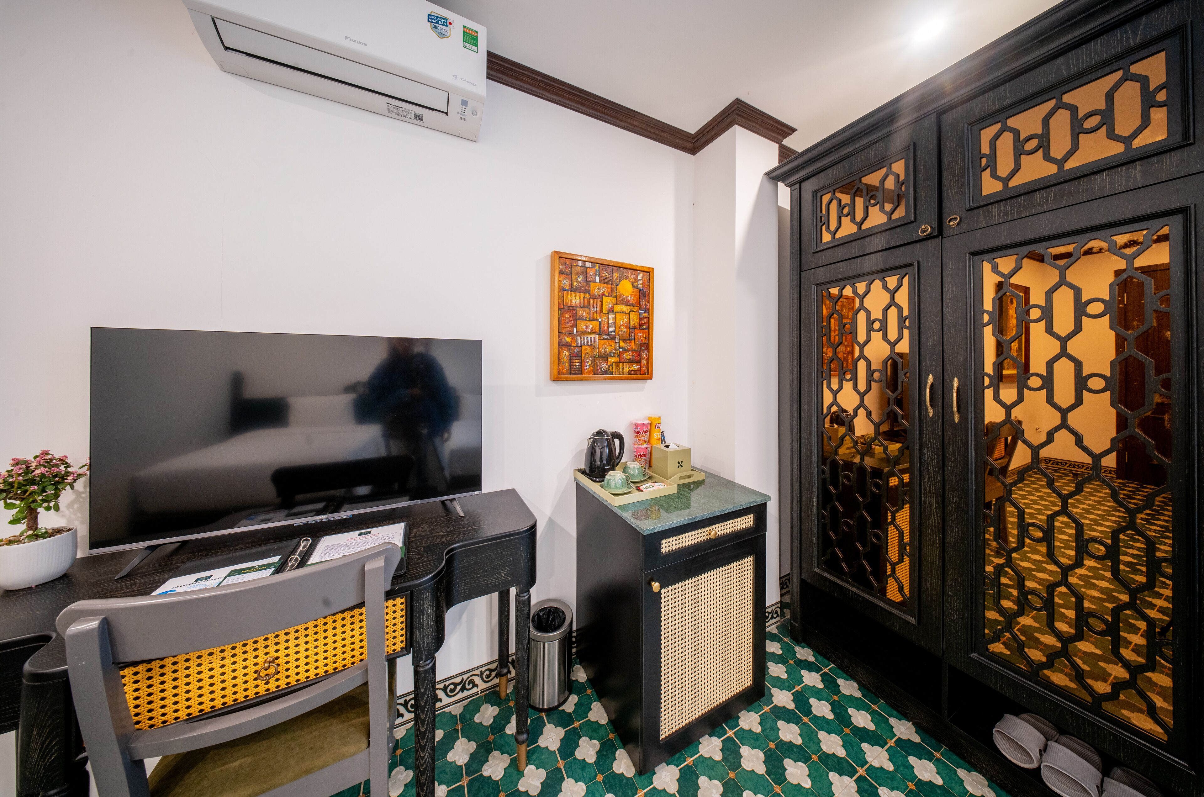 A room with a flat-screen TV, a small table with a kettle, a chair, a trash bin, and a decorative wall piece.