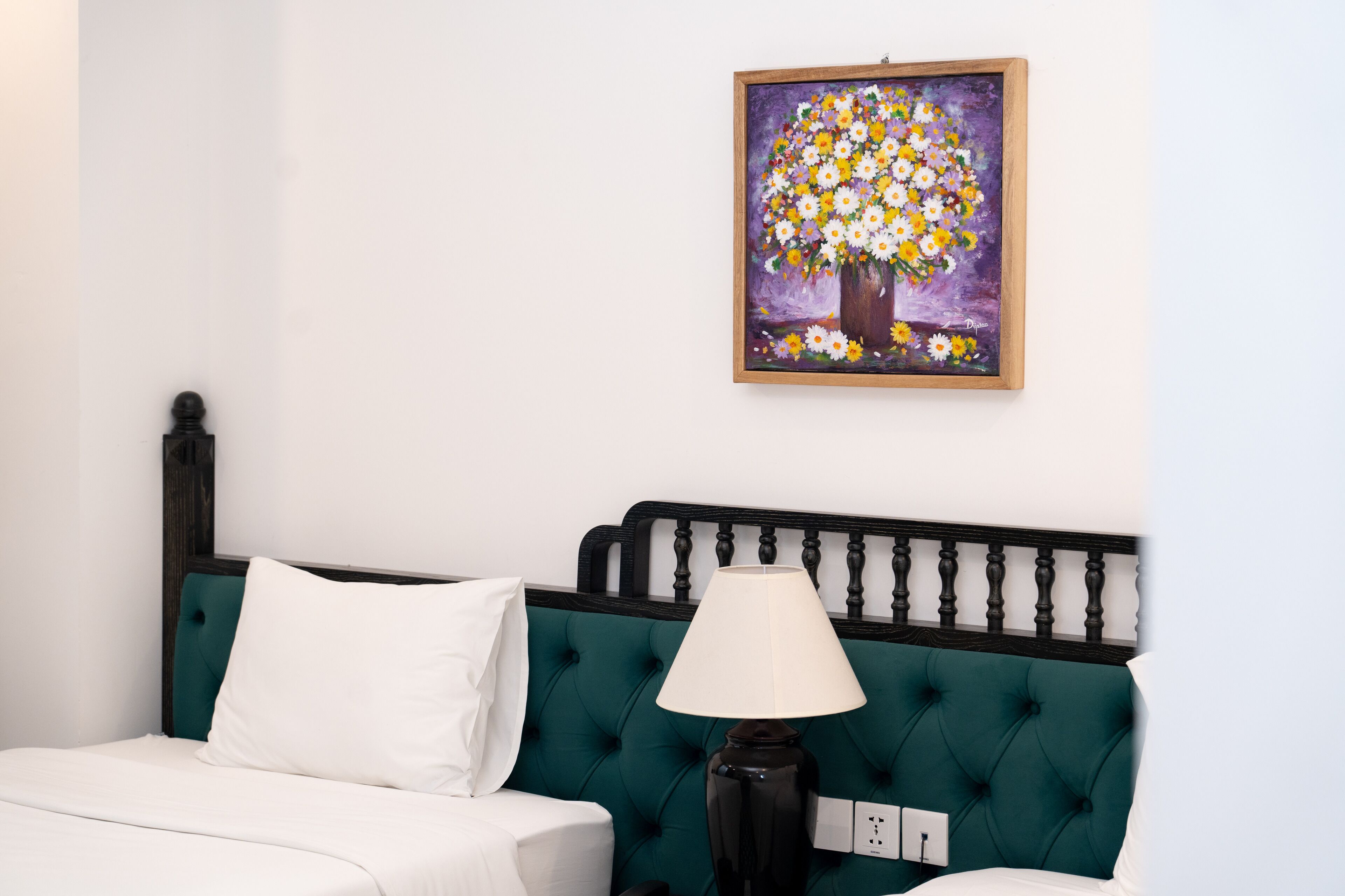A bedroom with a teal upholstered headboard, a white bed, a black lamp, and a painting of a tree with flowers on the wall.