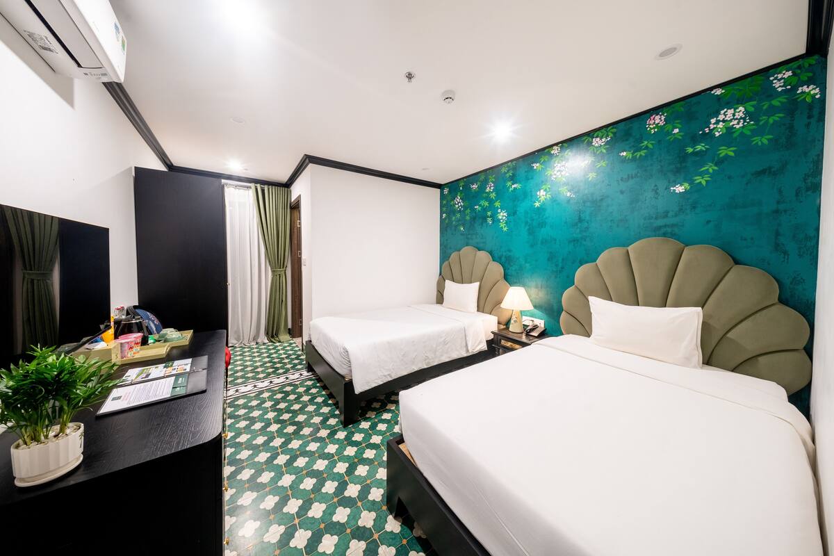 Habitación de hotel con dos camas, un escritorio con una planta, un espejo y una pared verde azulado con papel tapiz floral.