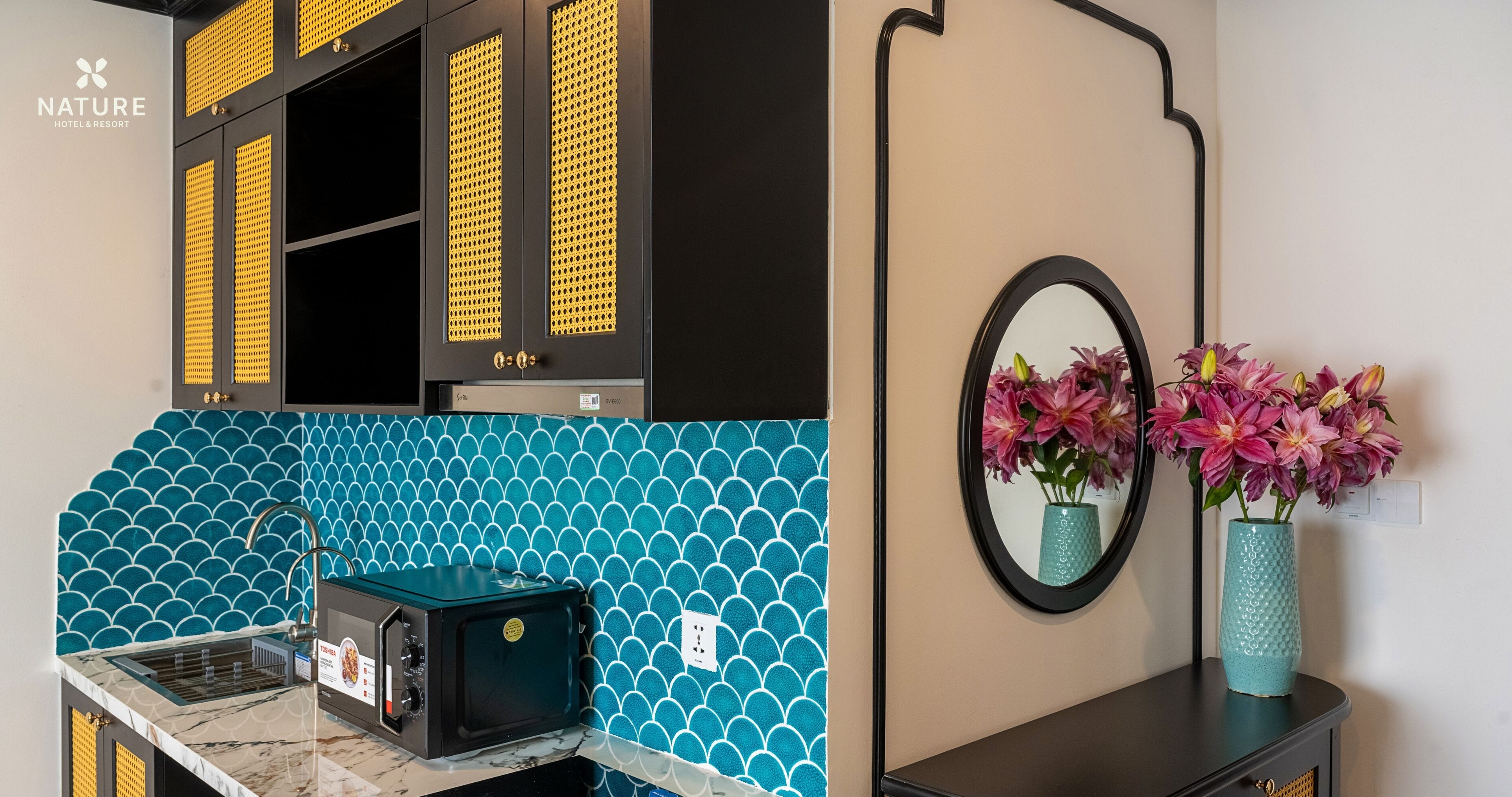 Una cocina con microondas, lavamanos y un salpicadero azul de azulejos. Hay un espejo con un florero sobre la encimera.