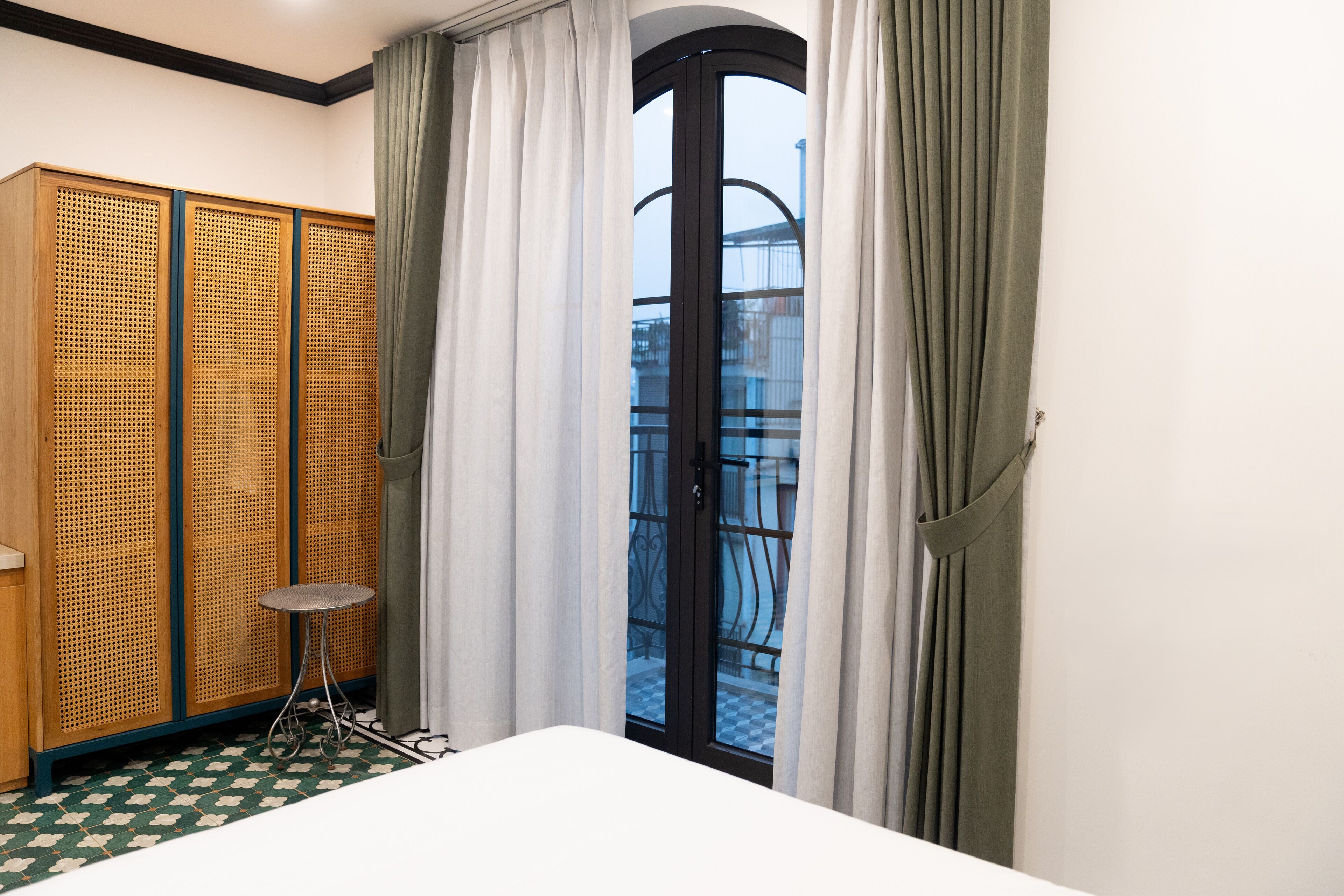 A hotel room with a bed, a small round table, a wooden wardrobe, and a balcony with a checkered floor.