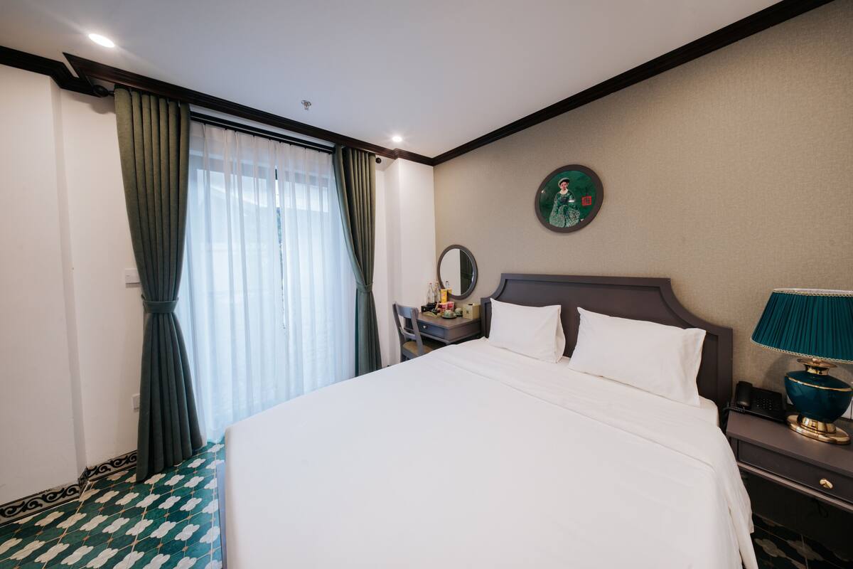 Habitación de hotel con una cama, un escritorio, una silla, una mesita de noche con lámpara, un reloj de pared y un ventanal con cortinas.