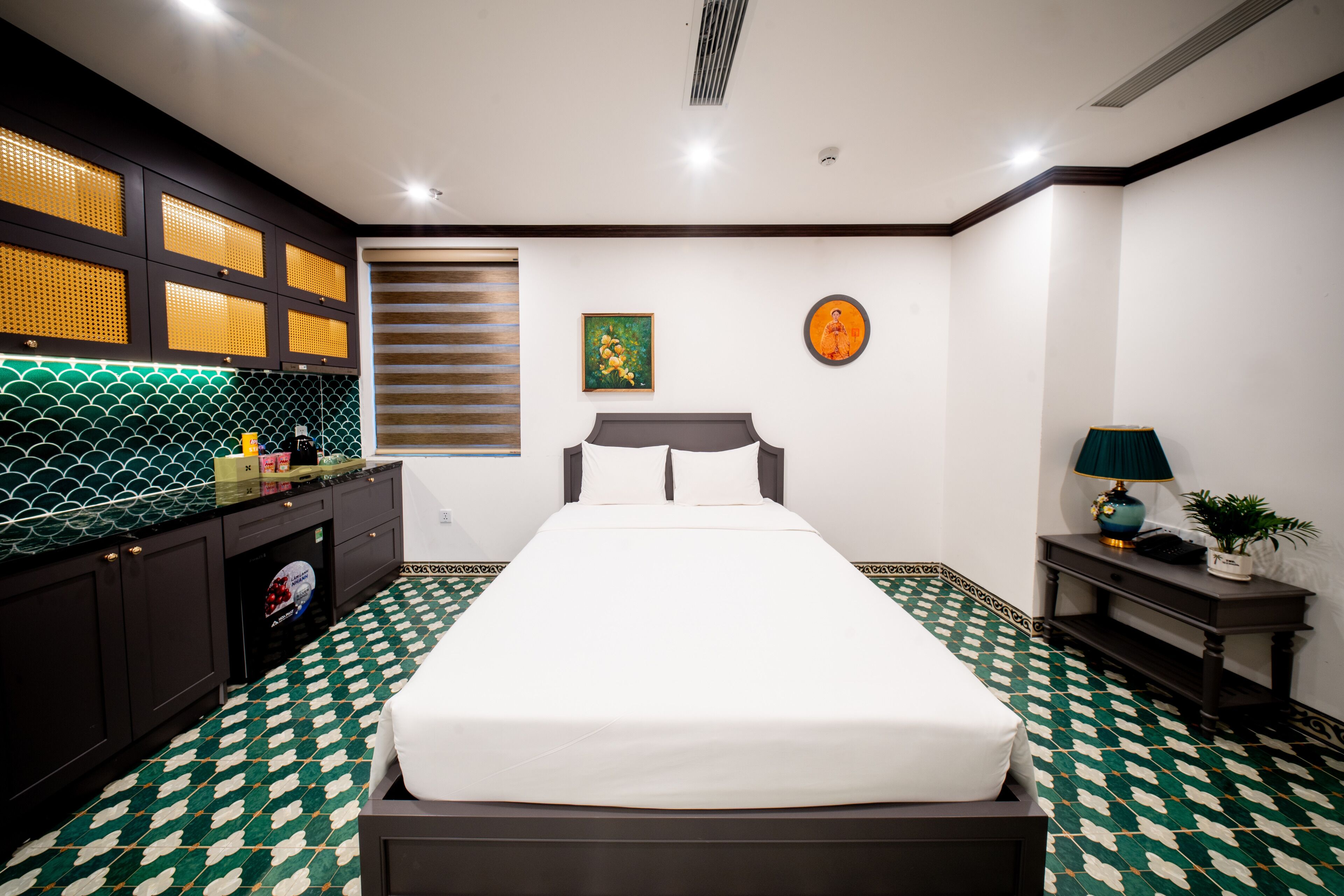 Habitació d'hotel amb llit, escriptori petit, tauleta de nit, finestra amb persianes i un objecte decoratiu a la paret.