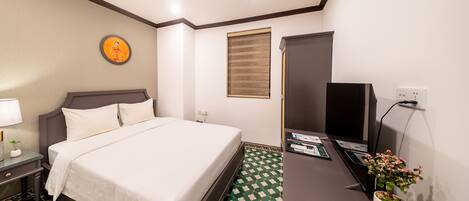 A hotel room with a bed, a desk with a computer, a TV, a plant, and a window with blinds.