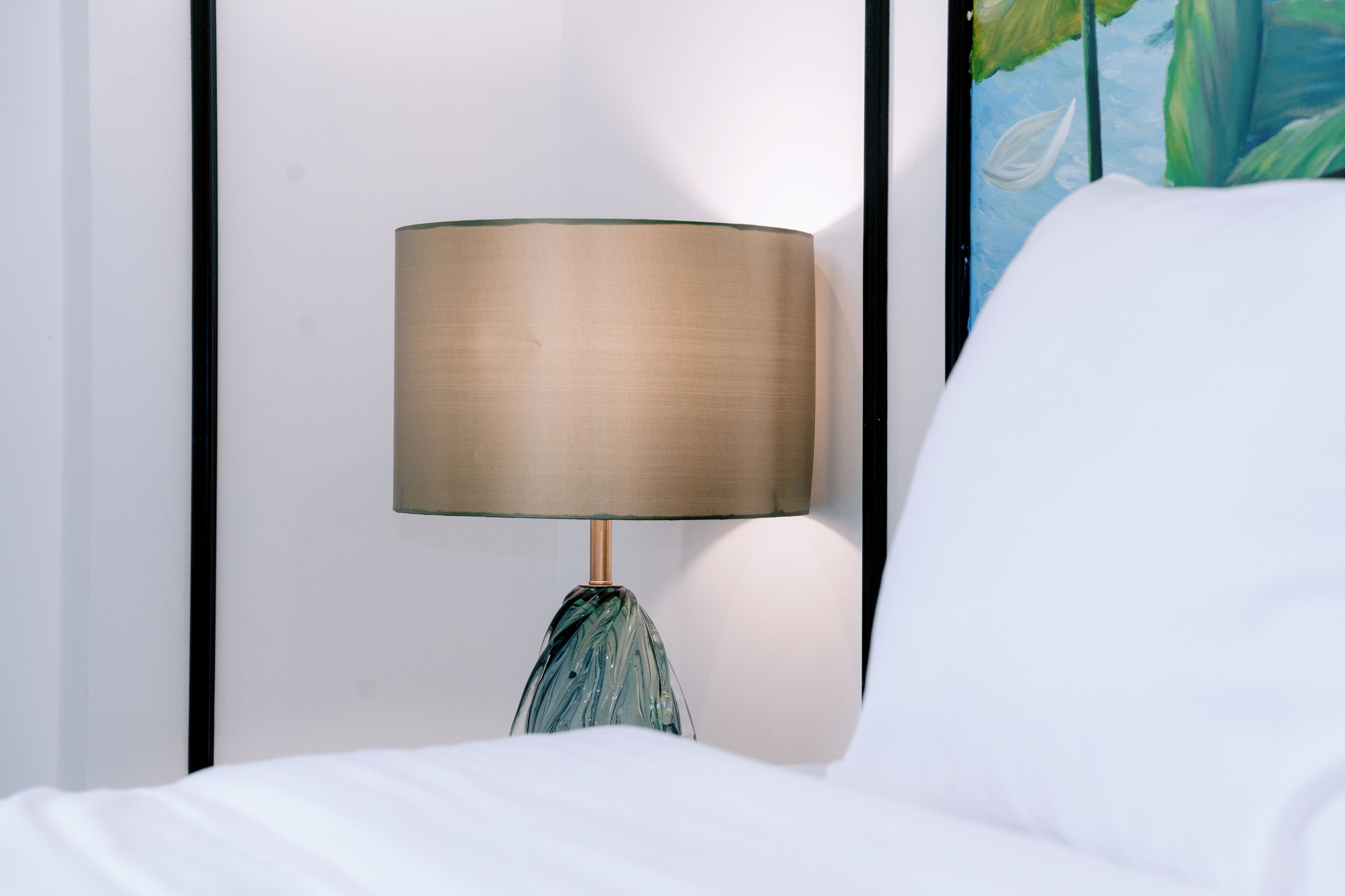 A bed with white sheets and a lamp with a beige shade and a blue base.