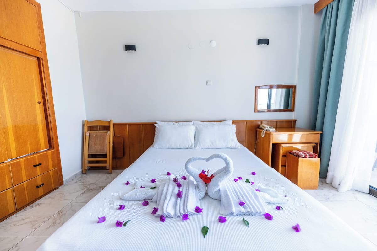 A hotel room with a neatly made bed, a wooden wardrobe, a small wooden chair, a bedside table with a lamp, and a mirror on the wall.