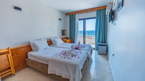 A hotel room with two beds, a wooden headboard, a nightstand, a small table with towels and flowers, a window with a view of the sea, and a wall-mounted air conditioning unit. - Çidihan Hotel (Kuşadası)