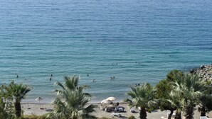 A beach with palm trees, a rocky shoreline, and people swimming in the water. - Çidihan Hotel (Kuşadası)