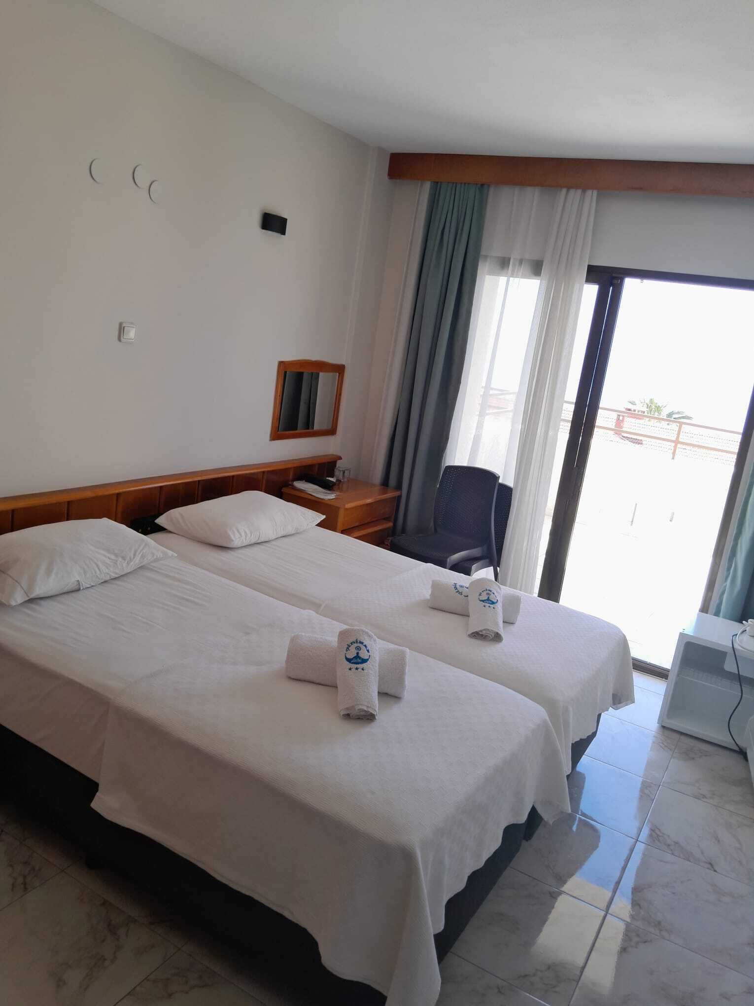 A hotel room with two beds, a wooden headboard, a nightstand, a chair, a window with curtains, and a view of the outside.