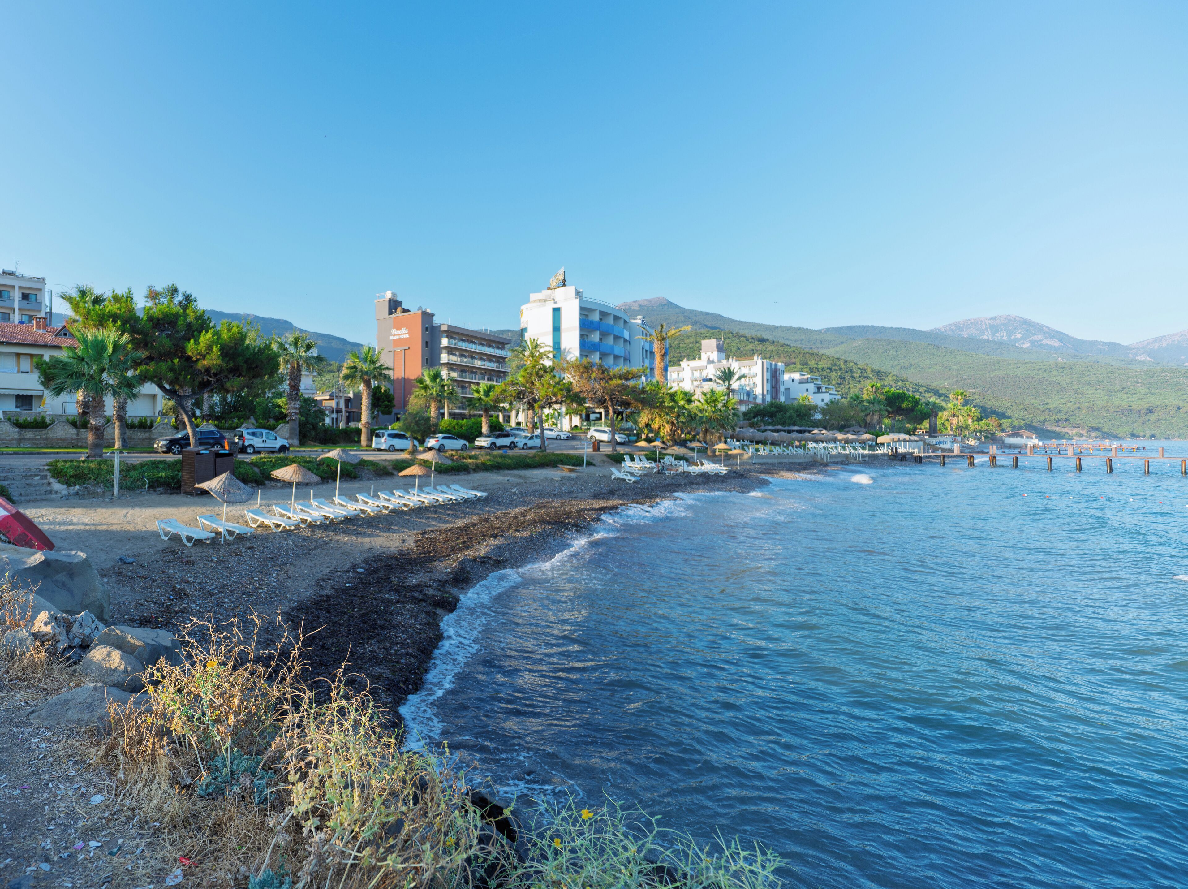 Une plage bordée d’une rangée de transats, un rivage rocheux et un hôtel en arrière-plan.
