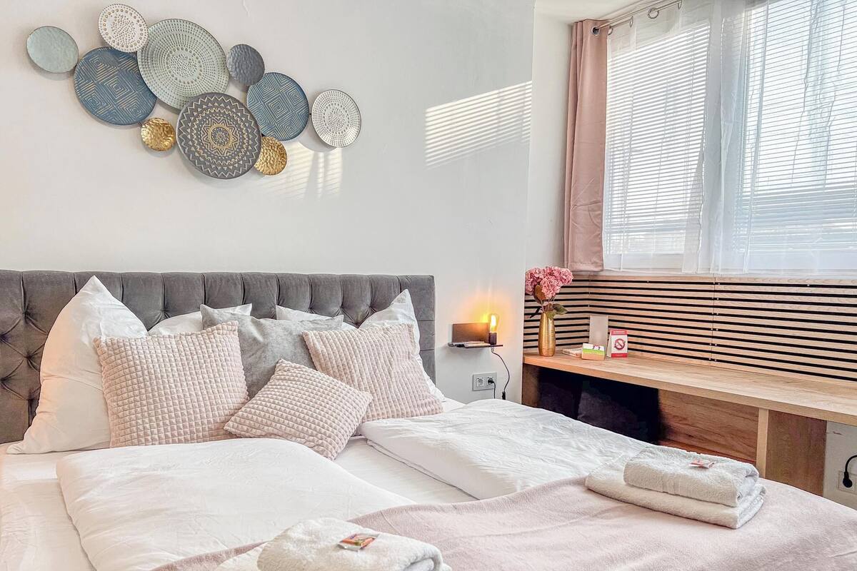 Una cama bien hecha con varias almohadas, una mesita de noche con una lámpara y un jarrón de flores.