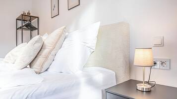 A neatly made bed with white linens and pillows, a bedside table with a lamp, and a wall-mounted power outlet.