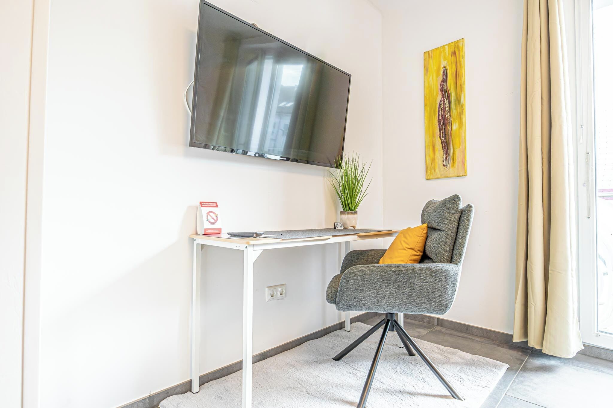 A modern room with a flat-screen TV mounted on the wall, a desk with a chair, a small potted plant, and a painting.