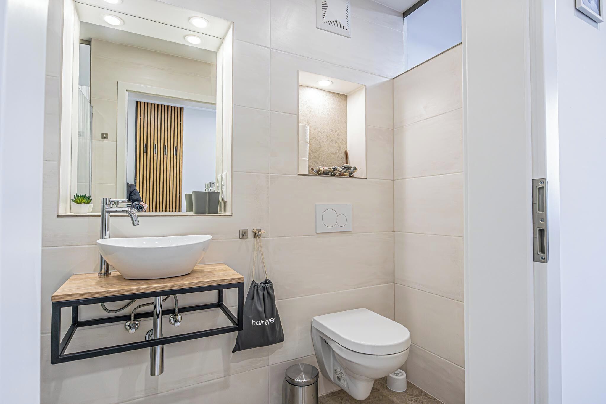 A modern bathroom with a large mirror, a vessel sink, a toilet, and a small shelf with toiletries.