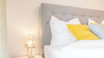Una cama bien hecha con cabecera capitonada, almohadas blancas y amarillas, y una lámpara de noche sobre una mesita blanca.