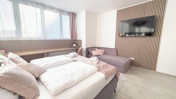 A modern bedroom with a bed, a sofa, a TV mounted on the wall, and a window with curtains.