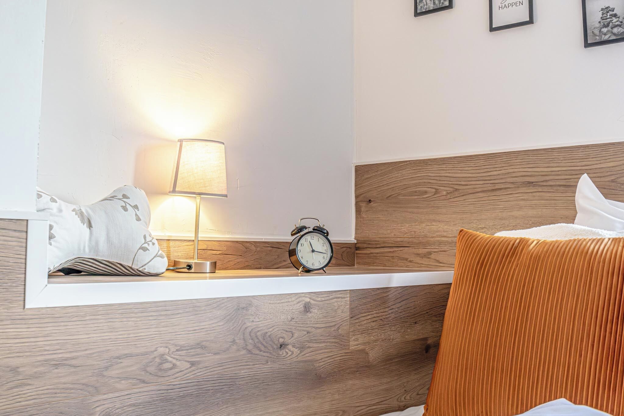 Habitación de hotel ordenada con cabecera de madera, mesita de noche con lámpara, reloj despertador y almohada.