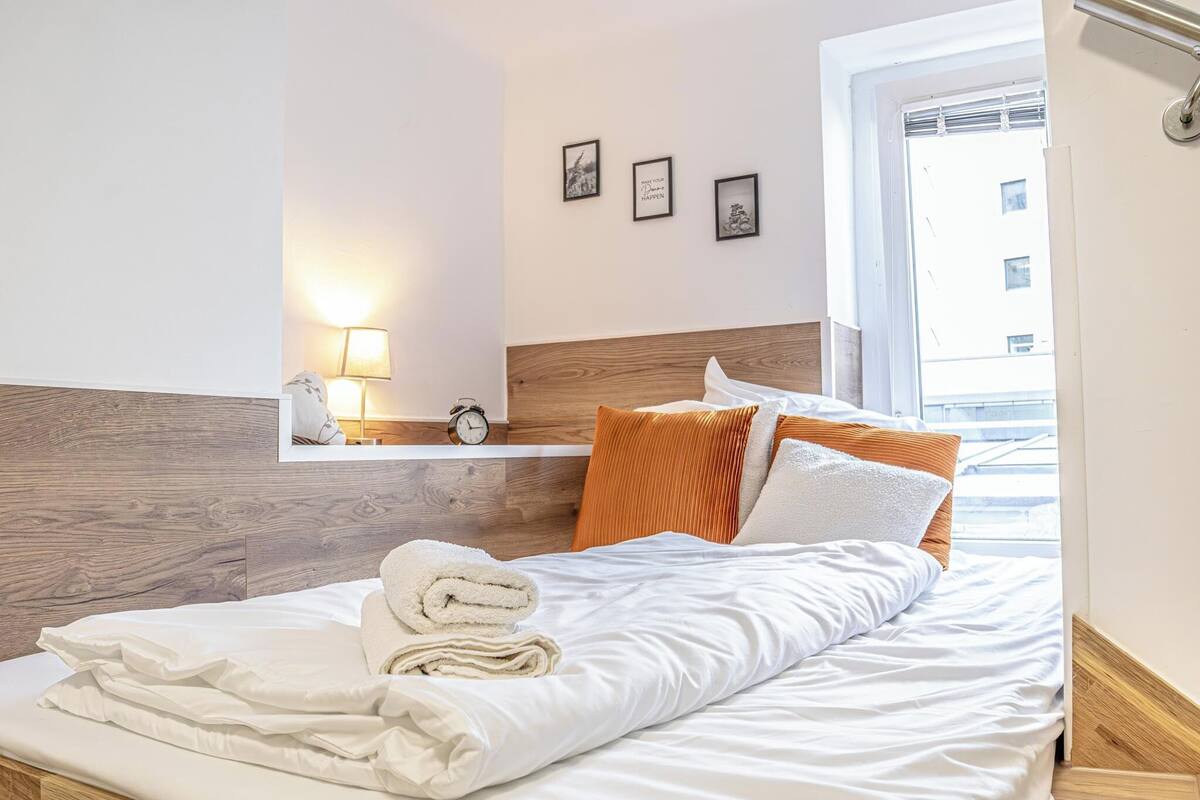Una cama bien tendida con sábanas blancas y una toalla doblada, cabecera de madera, lámpara de noche y ventana con vistas a edificios.