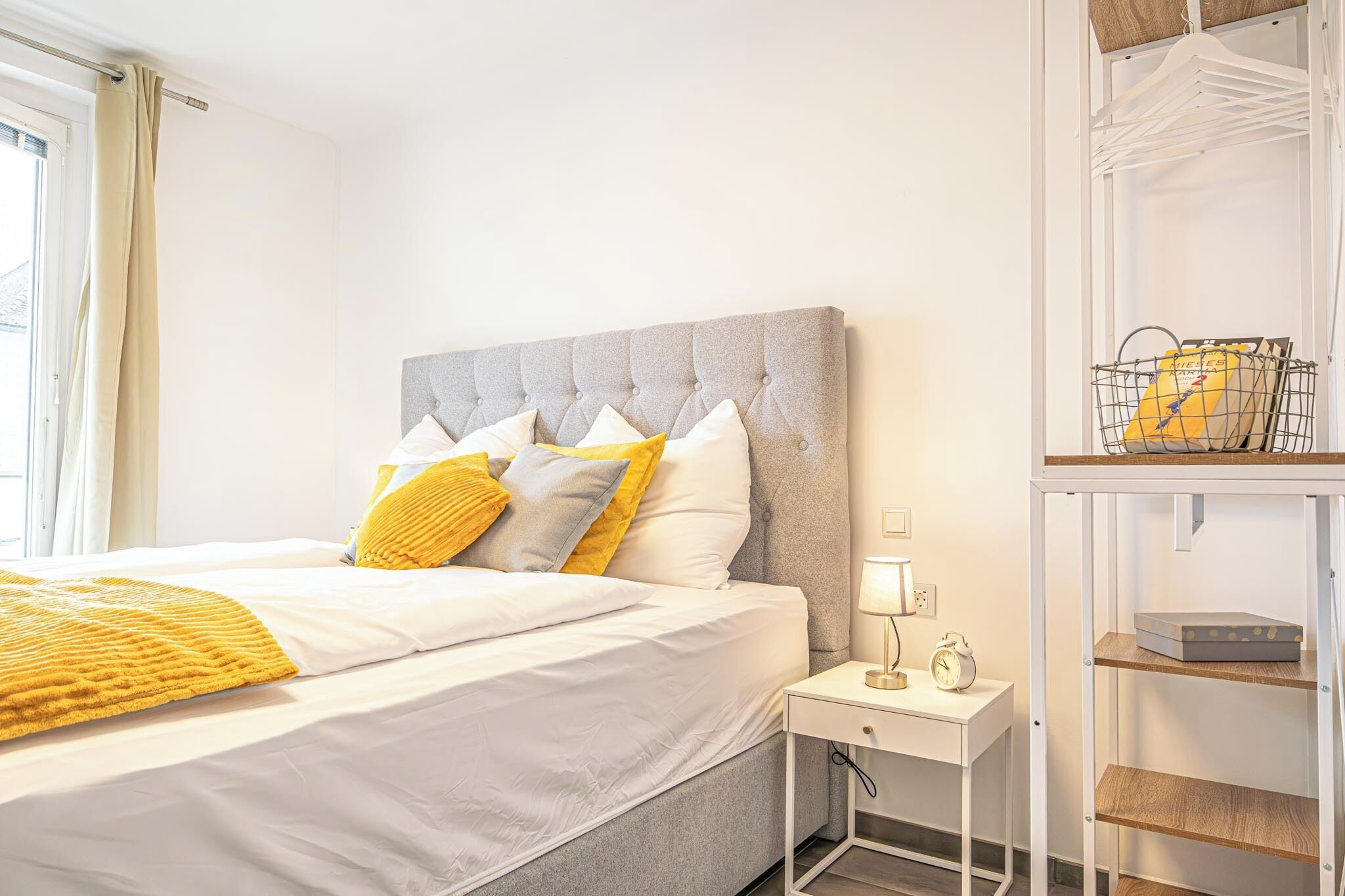 Una cama bien tendida con ropa de cama blanca y amarilla, un cabecero tapizado en gris y una mesita de noche con una lámpara y un despertador.
