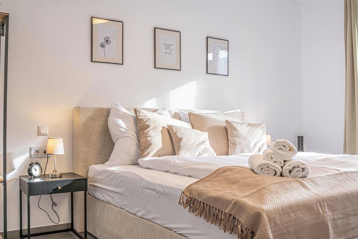 Una cama bien hecha con una manta color beige, una lámpara de noche y una mesita con un reloj.