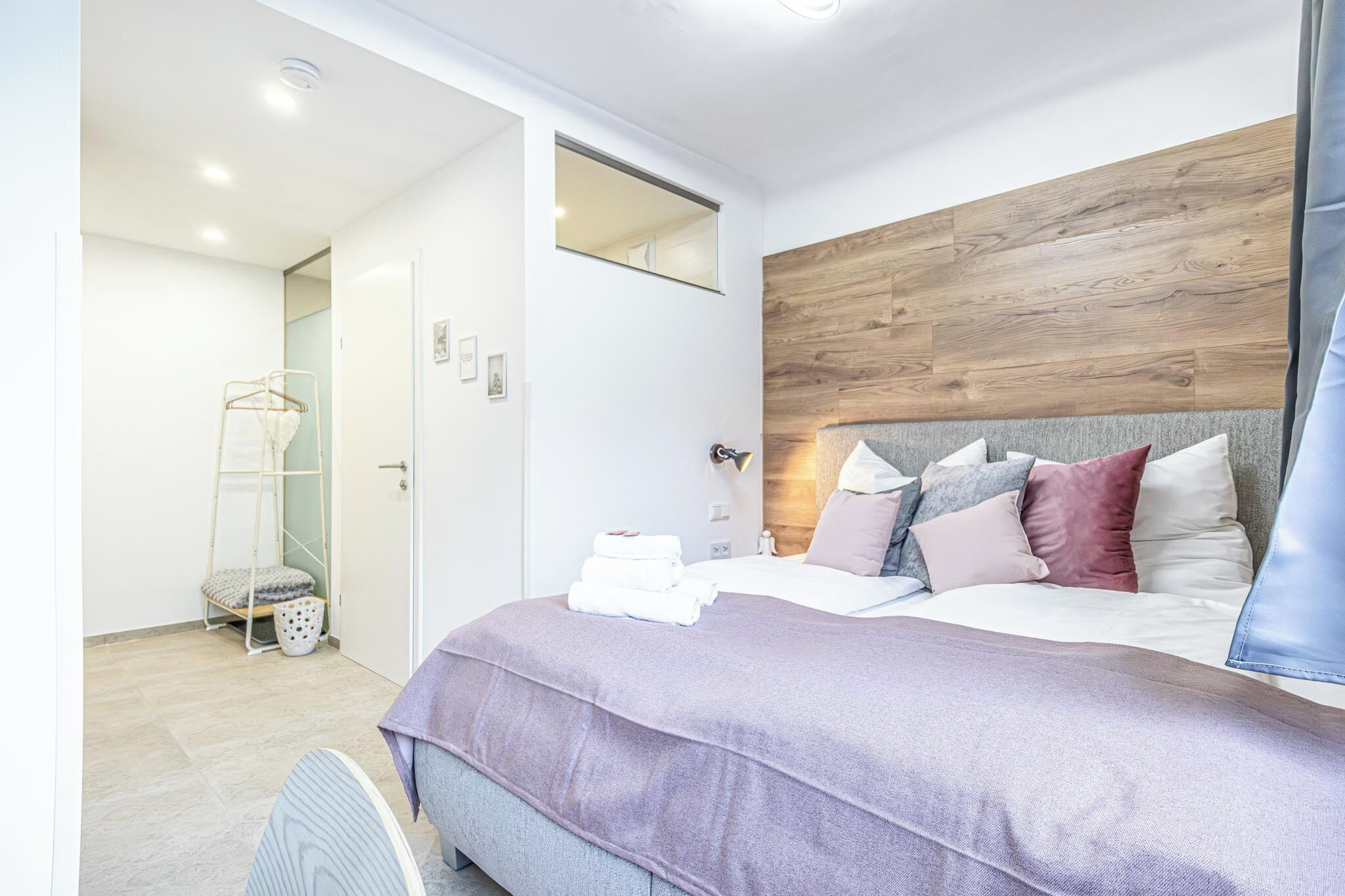 Quarto moderno com cabeceira de madeira, cama com v árias almofadas, mesa de cabeceira e um porta-toalhas.