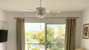 Un ampio soggiorno con ventilatore a soffitto, un divano beige, un tavolino da caffè con piano in vetro e un'ampia porta scorrevole in vetro che dà su un balcone con vista su vegetazione e una struttura blu.