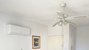 Un soggiorno con un divano color beige, un tavolino con piano in vetro, una ventola a soffitto e una porta.