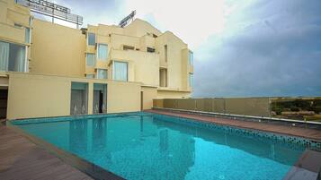 Piscina com deck de madeira, um edifício com várias janelas e céu nublado.