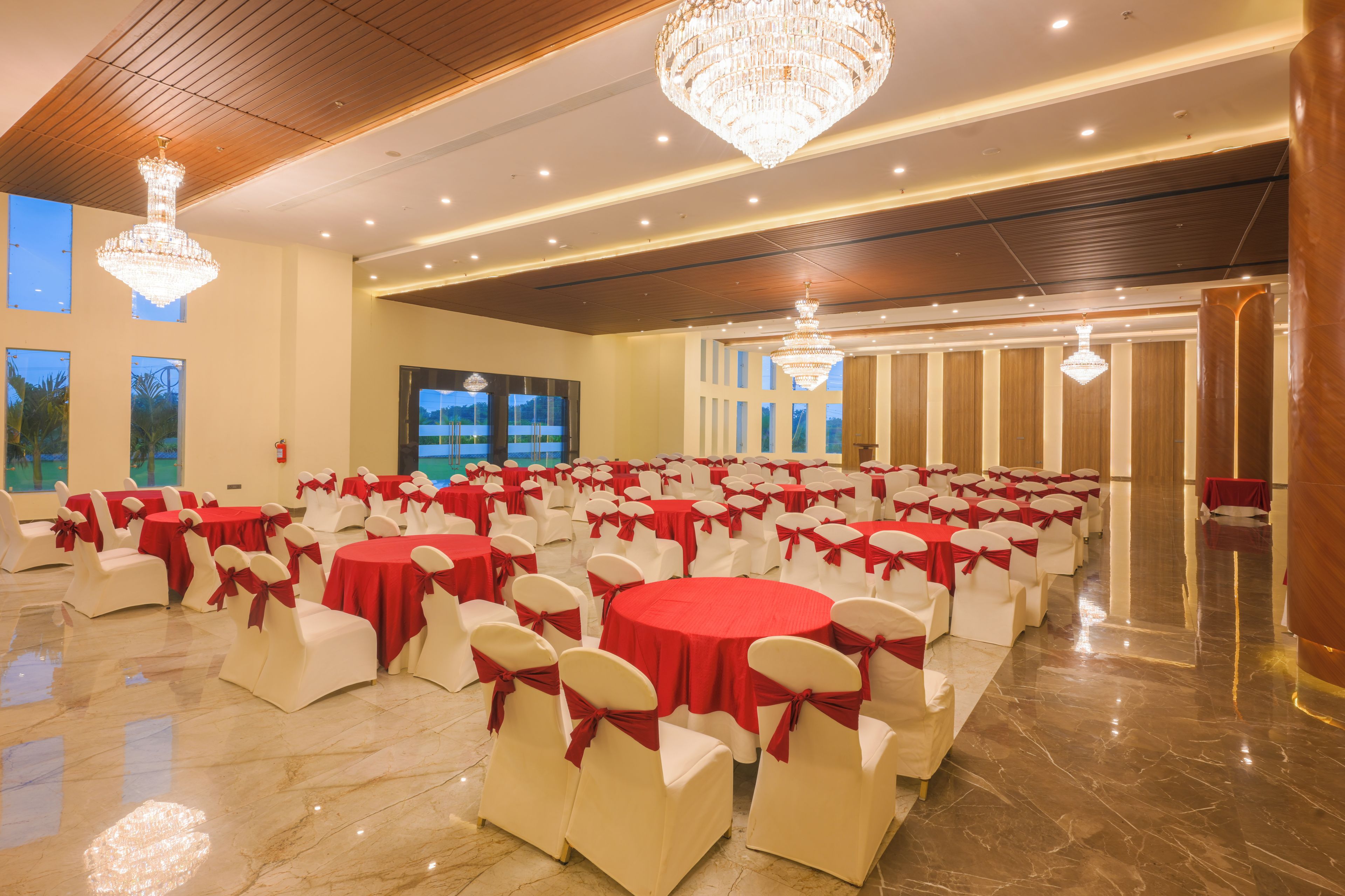 Un salón de banquetes con mesas redondas, sillas blancas con cintas rojas, candelabros y un ventanal con vistas.