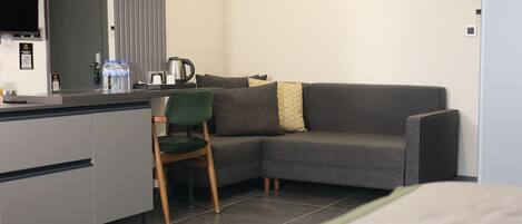 A modern hotel room with a grey sofa, a green chair, a small kitchen area with a kettle, and a bed.