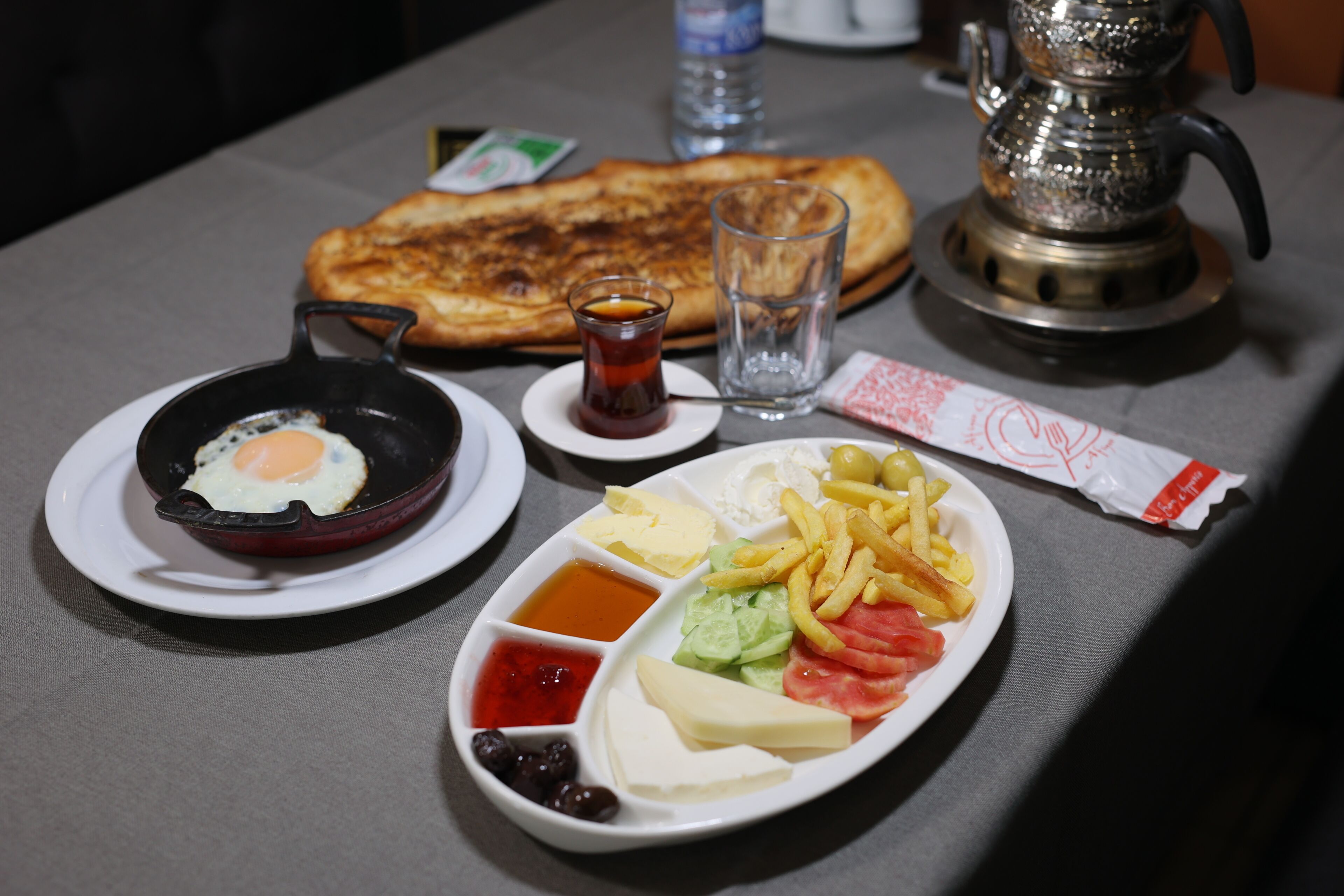 A table with a plate of food including eggs, cheese, tomatoes, lettuce, and olives; a glass of water; a small cup of tea; and a metal teapot.