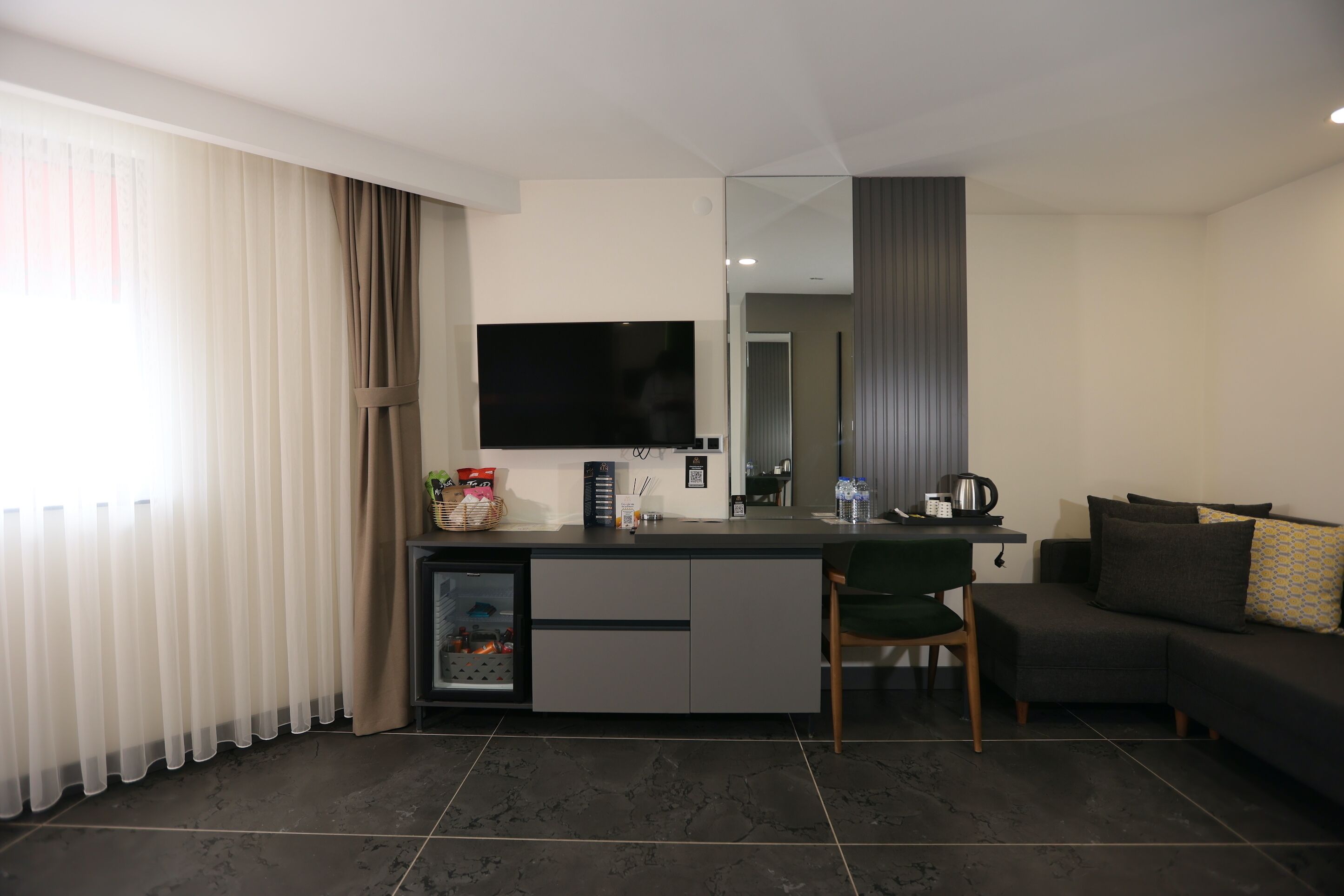Una sala de estar moderna con un sofá, una silla, un televisor de pantalla plana, una pequeña cocina y un gran ventanal con cortinas.