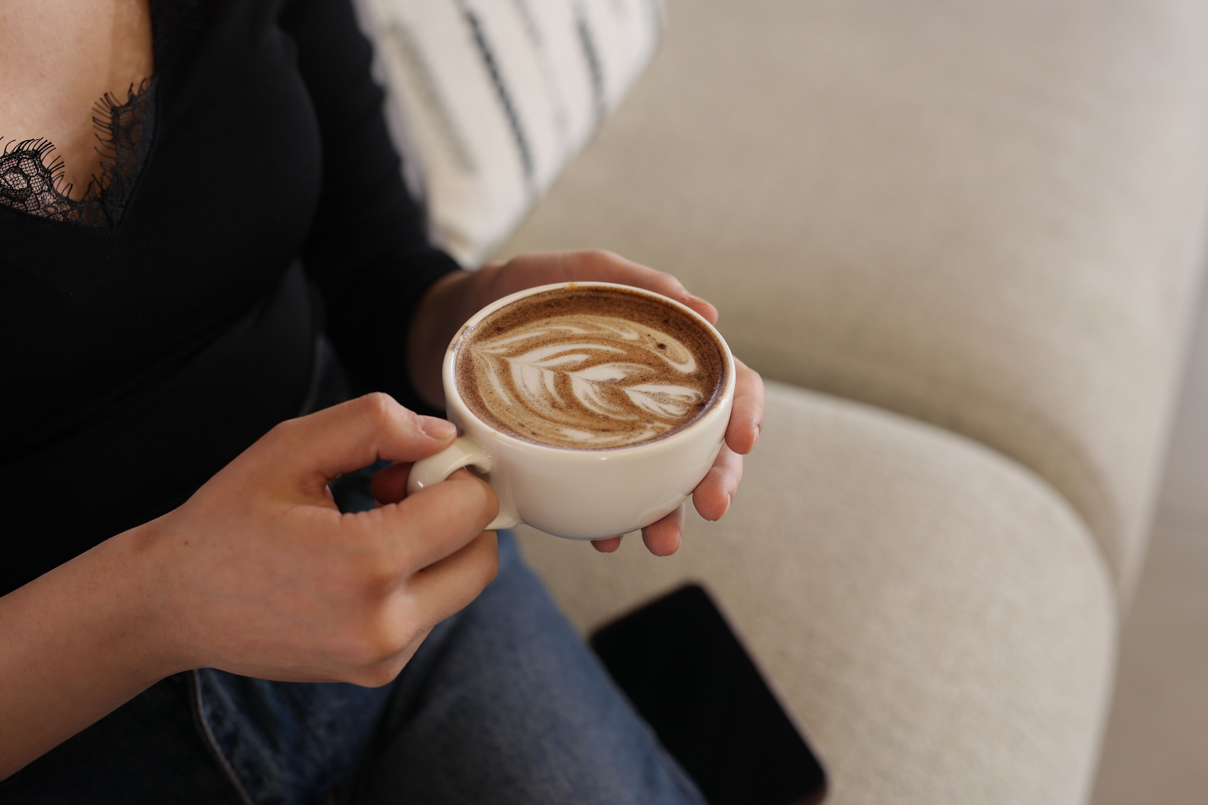 A person holding a cup of coffee with latte art, seated on a couch with a smartphone on the floor beside them.