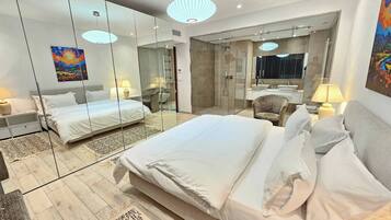 A modern hotel room with a large bed, a sitting area with a chair, a glass-enclosed shower, and a large mirror reflecting the room.