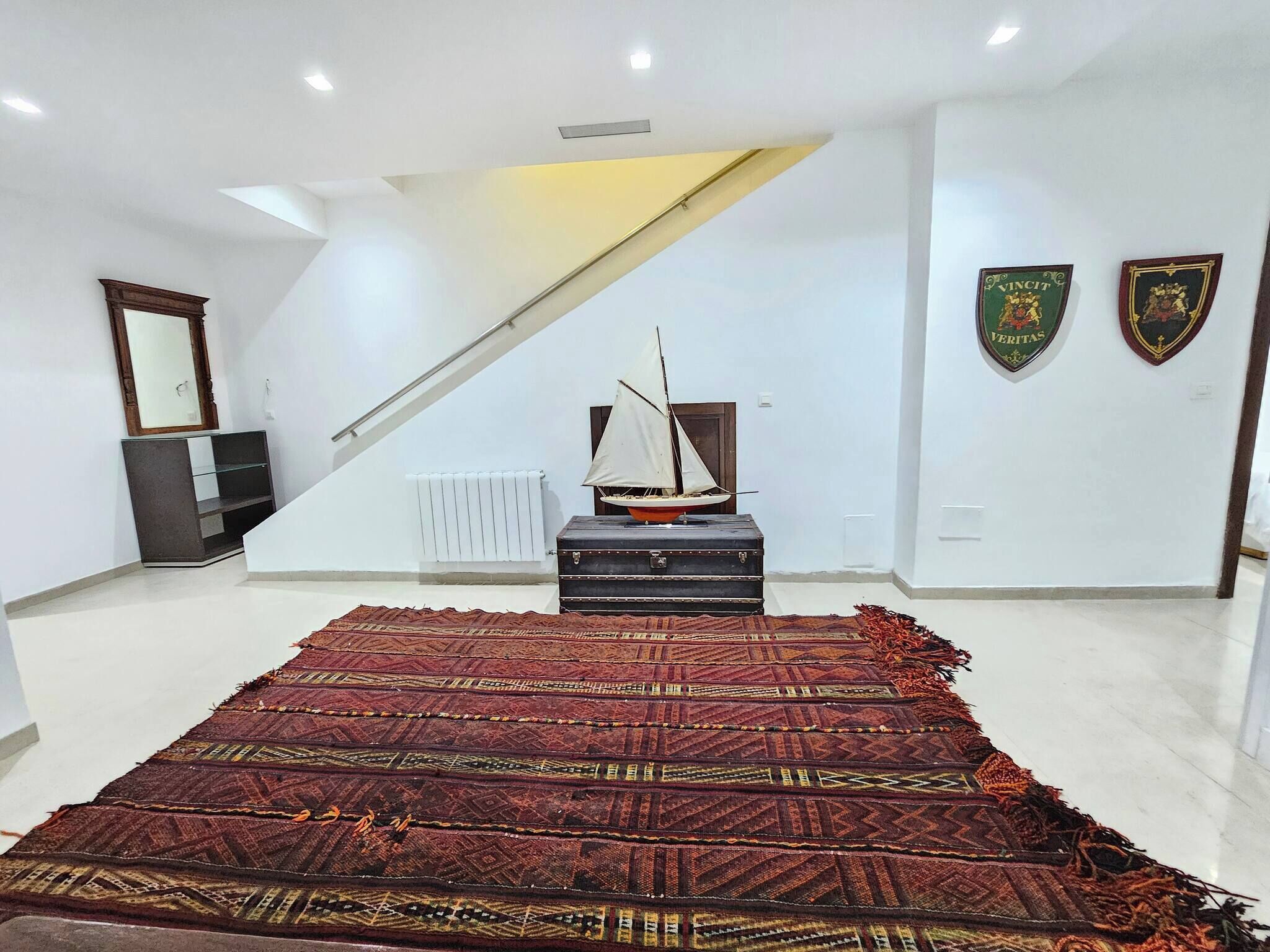 Una habitación amplia con un gran tapete estampado, un modelo de velero y dos escudos en la pared.