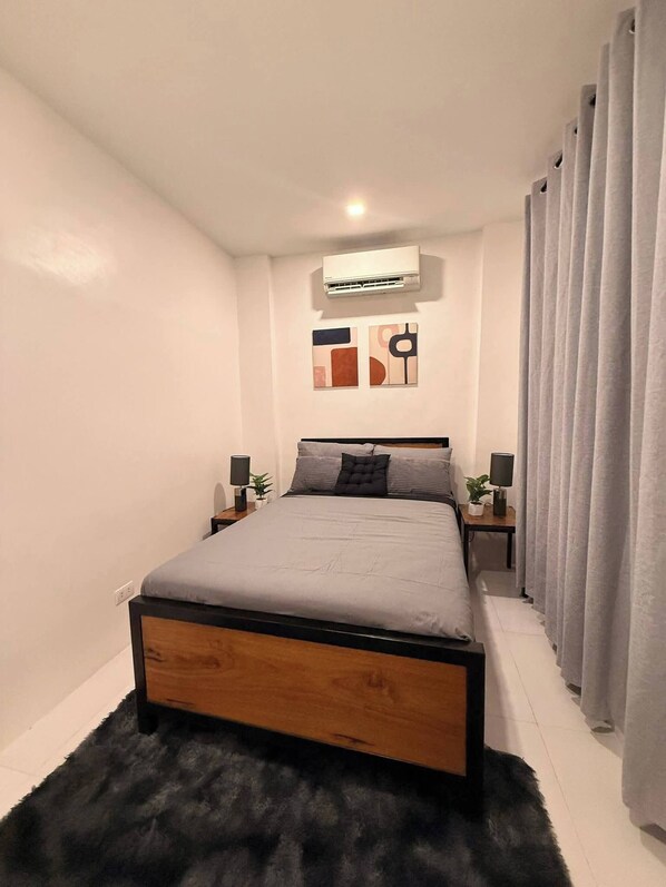 Une chambre moderne avec un cadre de lit en bois, un tapis moelleux, un climatiseur fixé au mur et deux tables de chevet ornées de lampes et de plantes.