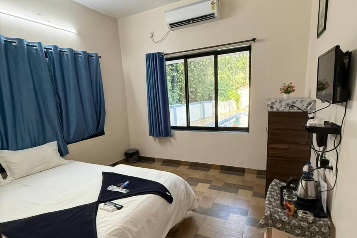 A hotel room with a bed, a ceiling fan, an air conditioning unit, a window with curtains, a small desk with a kettle, and a television.