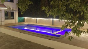 Una piscina rectangular con iluminación azul, rodeada por un muro bajo y un árbol.