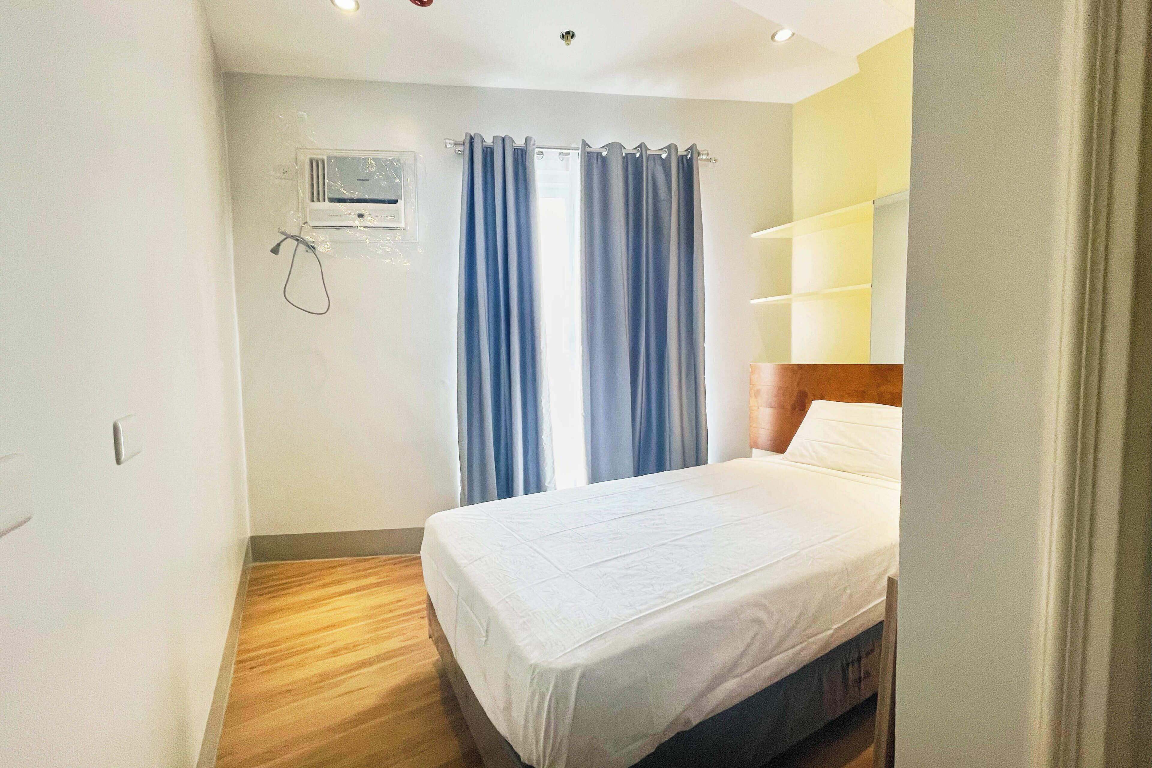 A hotel room with a bed, a window with curtains, a wall-mounted air conditioning unit, and a wooden floor.